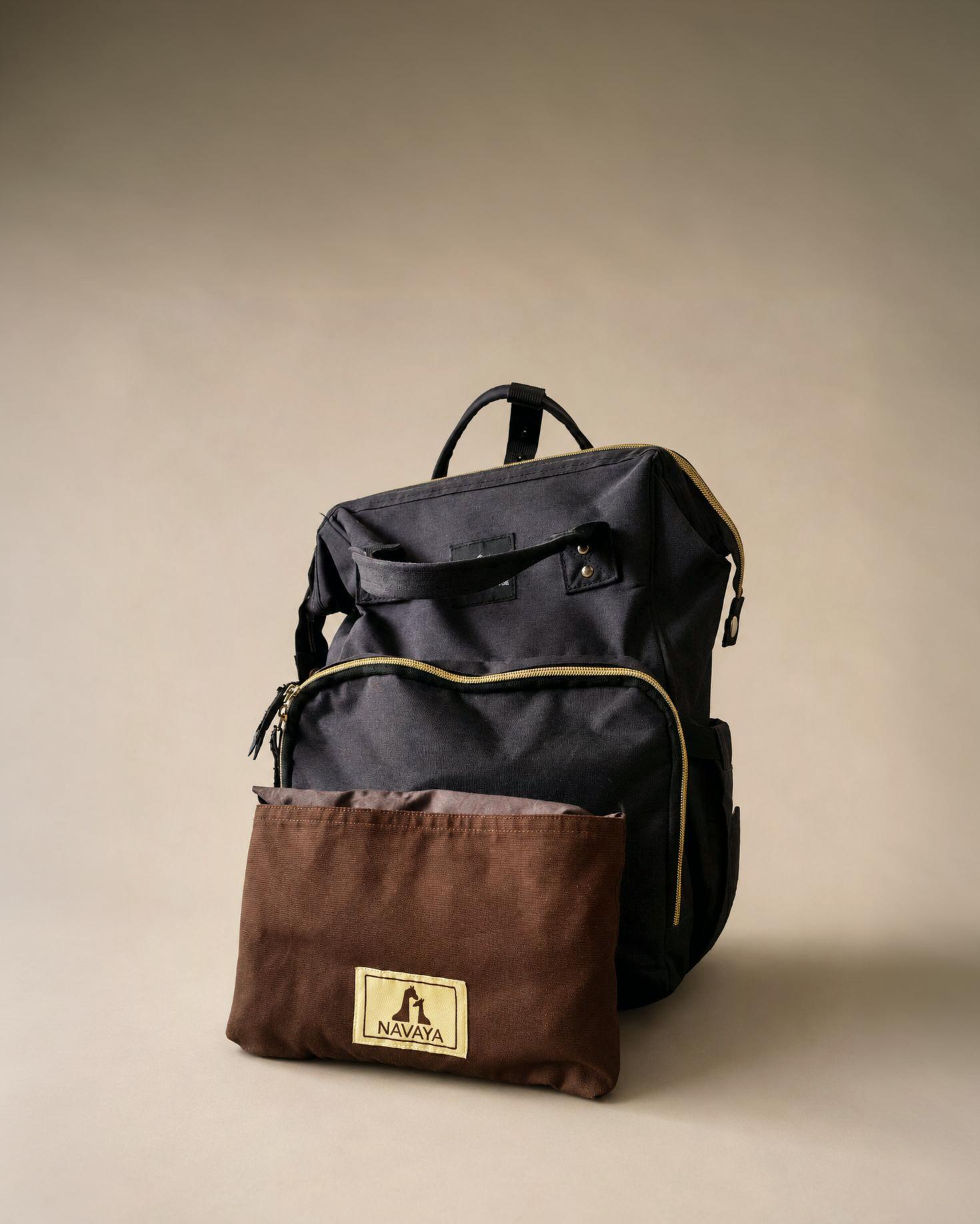 A black canvas backpack with gold-toned zipper trim stands against a warm beige studio background, with a brown pouch in front featuring a visible NINJA logo patch.